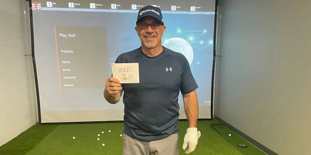 A golfer holding up a handwritten HOLE IN ONE card in front of the simulator screen