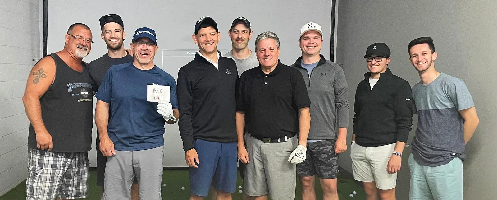Nine golfers lined up in the simulator bay after a round, one holding a handwritten card that reads HOLE IN ONE!!!