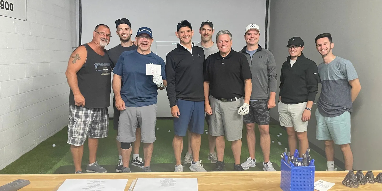 Nine golfers lined up in the simulator bay after a round, one holding a card that reads HOLE IN ONE!!!