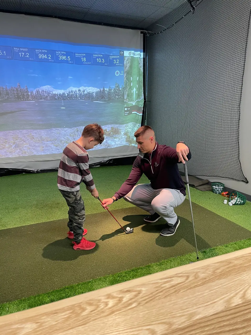 A coach crouching beside a young student, setting up the ball on the hitting mat